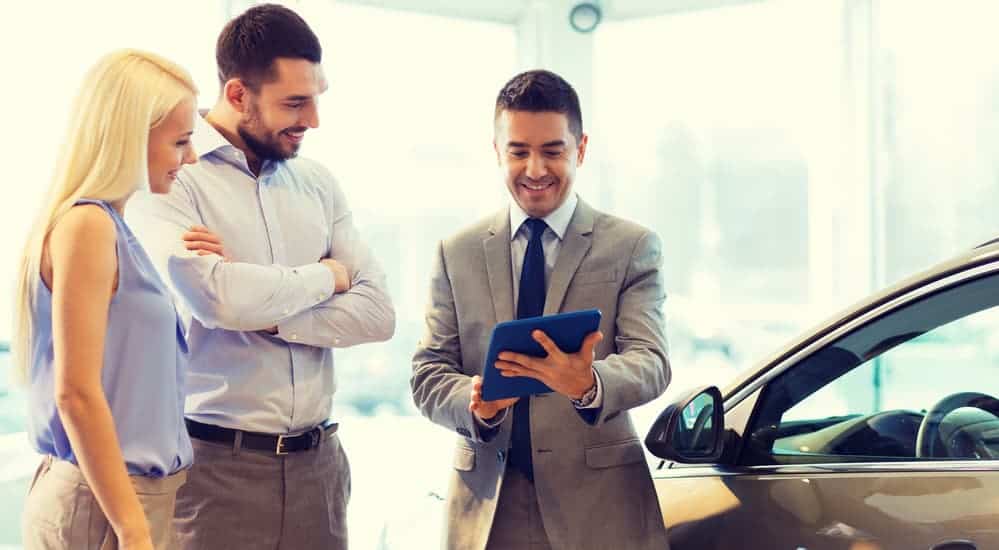 A smiling car salesman is using his iPad to help show smiling customer car information. 