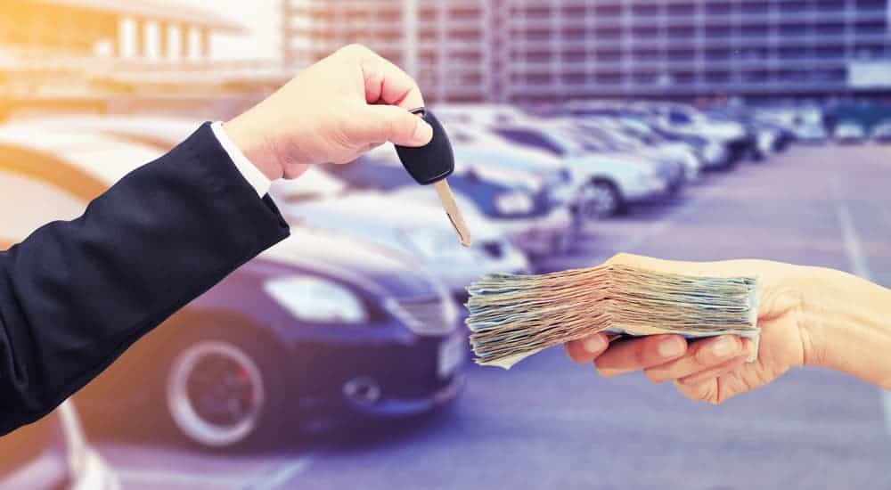 A hand is holding a car key while another hand is holding money with a blurry used car dealerships in the background. 