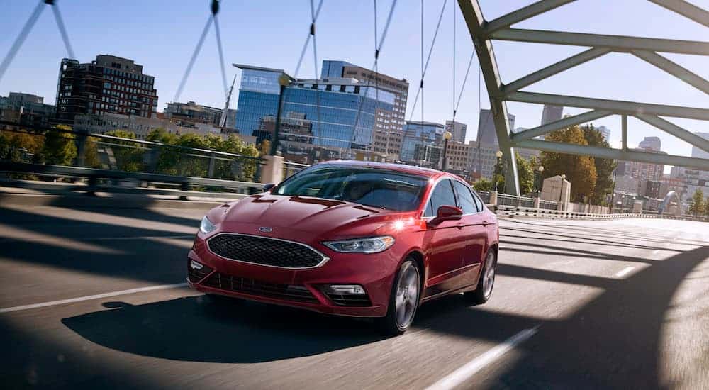 A red certified pre-owned 2017 Ford Fusion is driving on a bridge with a city skyline in the distance. 