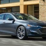 A blue 2020 Chevy Malibu is parked in front of a brick building with 2 men walking by it.