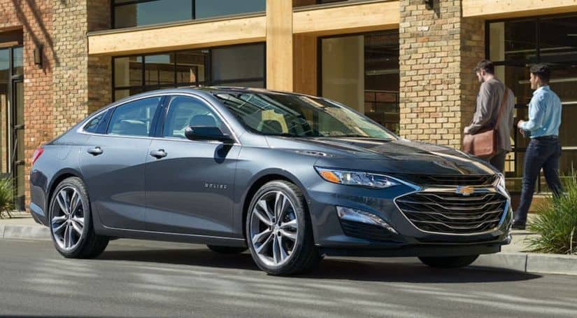 A blue 2020 Chevy Malibu is parked in front of a brick building with 2 men walking by it.