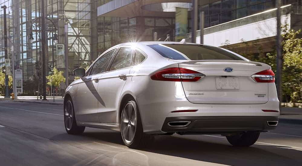 A white 2020 Ford Fusion is shown from the rear driving on a city street passed glass buildings. 