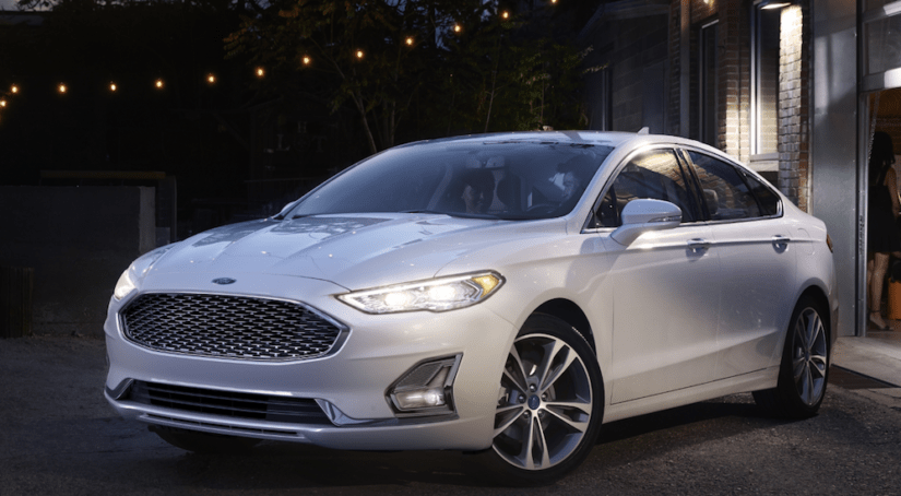 A white 2020 Ford Fusion is parked next to a brick building under lit string lights at night.