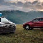Two 2020 Jeep Cherokee's, red and grey, are parked in a grassy field at dusk with mountains in the back.