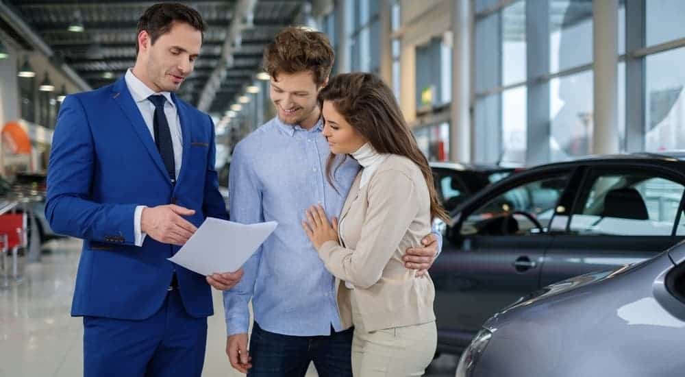 A smiling salesman is looking over a used car Carfax history report with a smiling couple at a dealership
