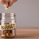 Fingers are shown putting a penny inside a jar that says "budget" on it.