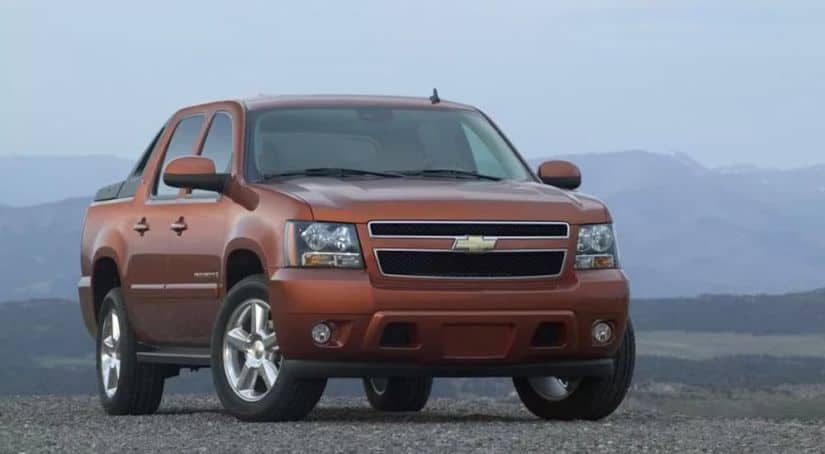 An orange 2011 Chevrolet Avalanche is parked on a gravel parking lot with mountains in the distance.