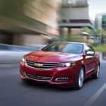 A red 2020 Chevy Impala is driving on a city street past blurry buildings.