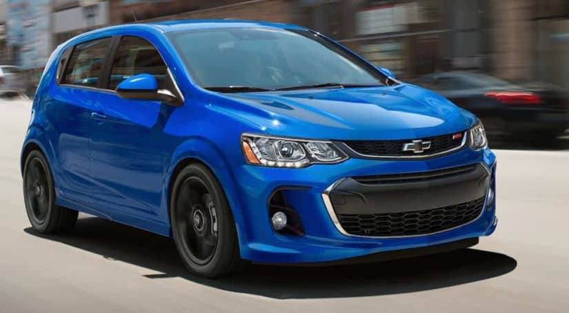 A blue 2020 Chevy Sonic is driving on a city street past blurred building.