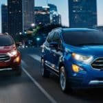 Two 2020 Ford Ecosports, one blue and one red, are driving on a city street at dusk.