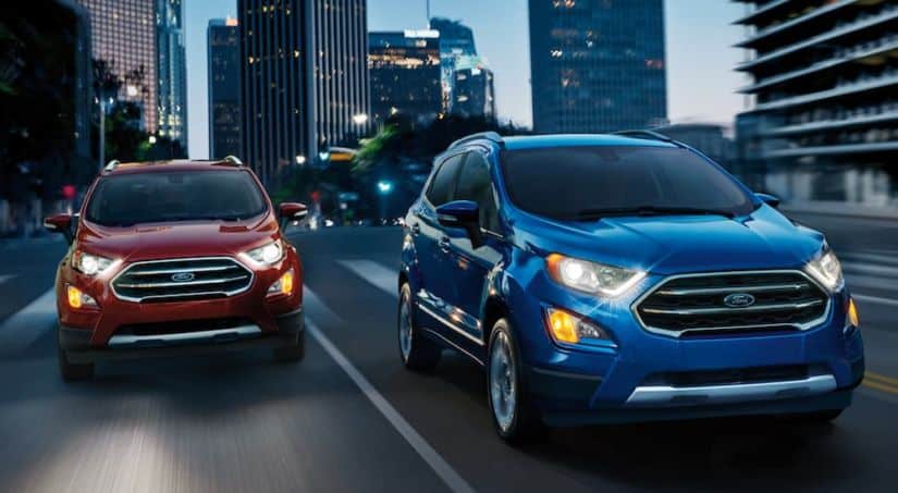 Two 2020 Ford Ecosports, one blue and one red, are driving on a city street at dusk.