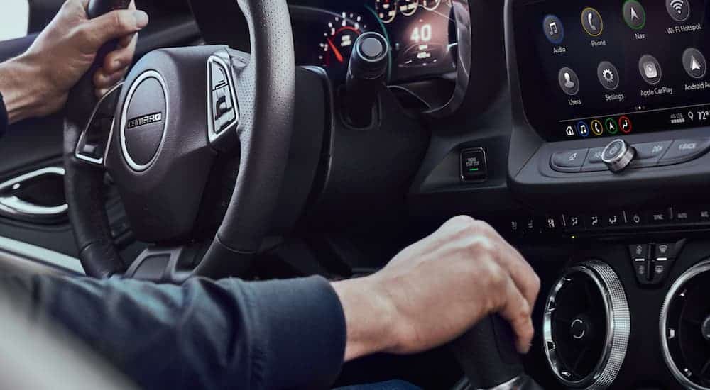 A close up is showing a man with one hand on the wheel and one on the shift knob of a 2021 Chevy Camaro.