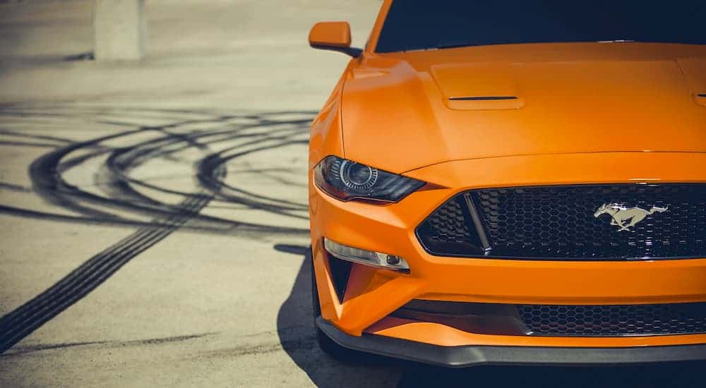 A close up is showing the front grille and headlights of an orange 2021 Ford Mustang with tire marks next to it.