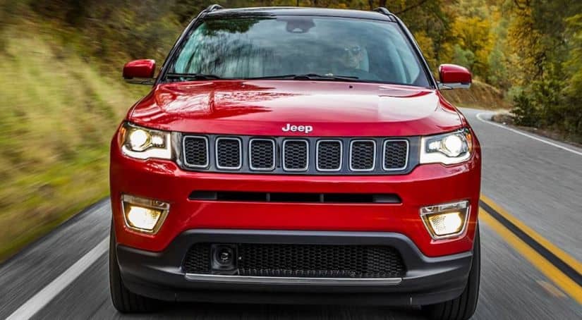 A red 2020 Jeep Compass is driving on a treelined road while facing forward.