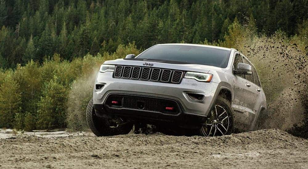 A grey 2020 Jeep Grand Cherokee Trailhawk is driving up a dirt hill with pine trees in the distance. 