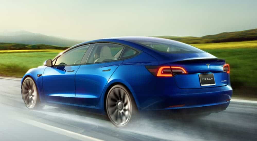 A blue 2021 Tesla Model 3 is shown from behind driving down a wet road.