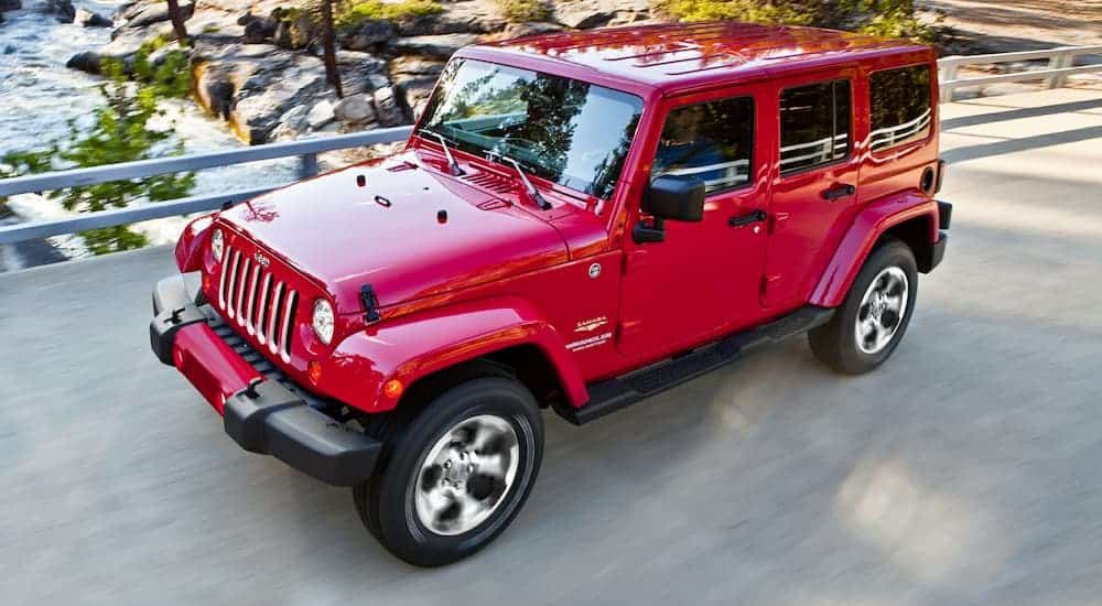 A red 2017 Jeep Wrangler Sahara, which is popular among used Jeeps for sale, is driving on a bridge over a river.