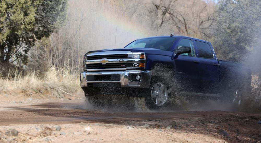 A blue 2016 Chevy Silverado 2500HD, which is the most popular option among used trucks, is driving through a large puddle in the woods.