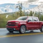 A red 2020 Chevy Silverado 1500 is driving on a wet road with snow covered mountains in the distance.
