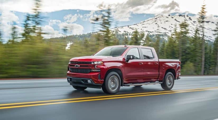 A red 2020 Chevy Silverado 1500 is driving on a wet road with snow covered mountains in the distance.
