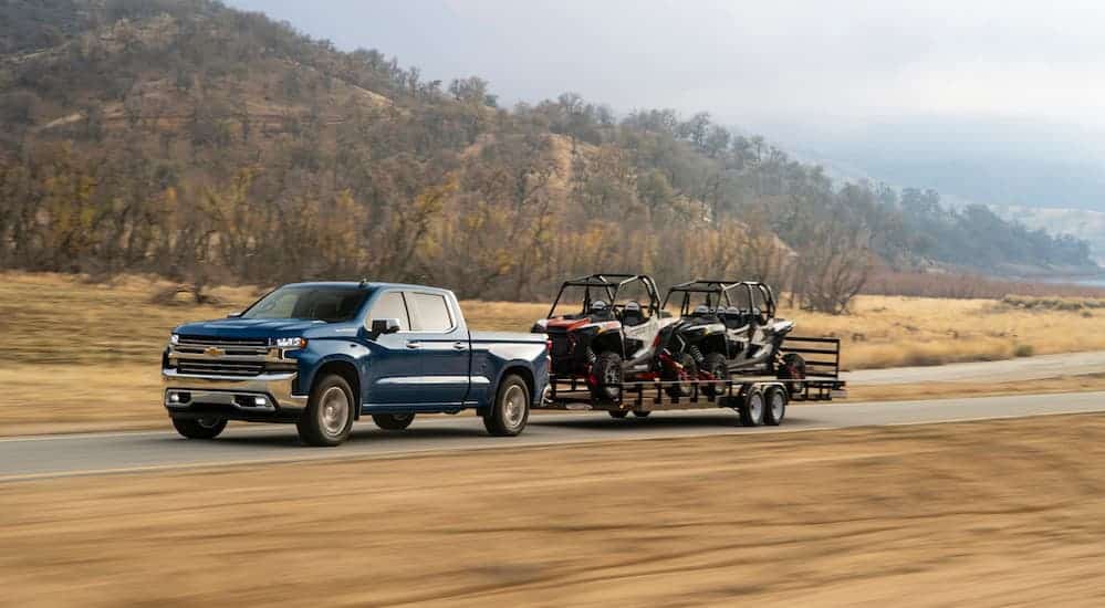 A blue 2020 Chevy Silverado 1500, which wins when comparing the 2020 Chevy Silverado vs 2020 Ram 1500, is towing two side by sides on a large trailer.