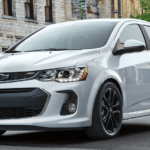A silver 2020 Chevy Sonic is driving on a city street past restaurants.