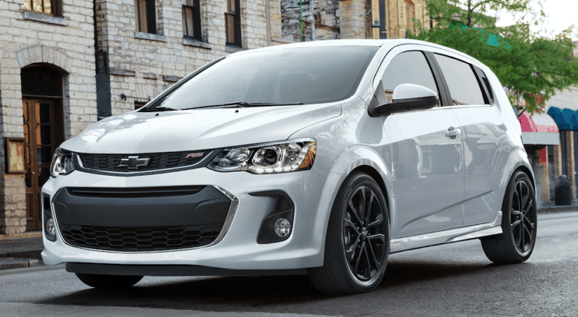 A silver 2020 Chevy Sonic is driving on a city street past restaurants.