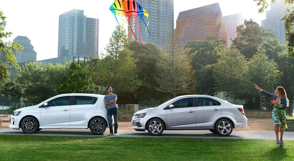 A man is watching his girlfriend fly kites while standing next to two 2020 Chevy Sonics in a park. 