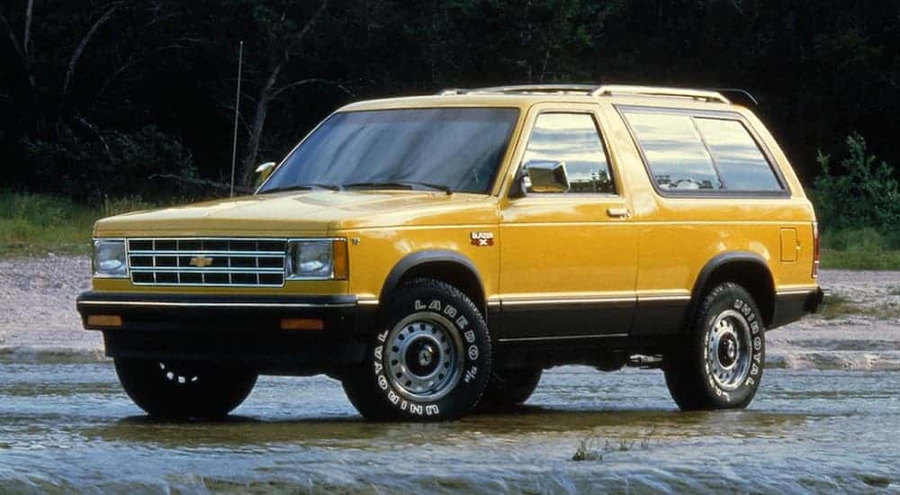 A yellow 1983 Chevy S10 Blazer is parked in front of dark trees.