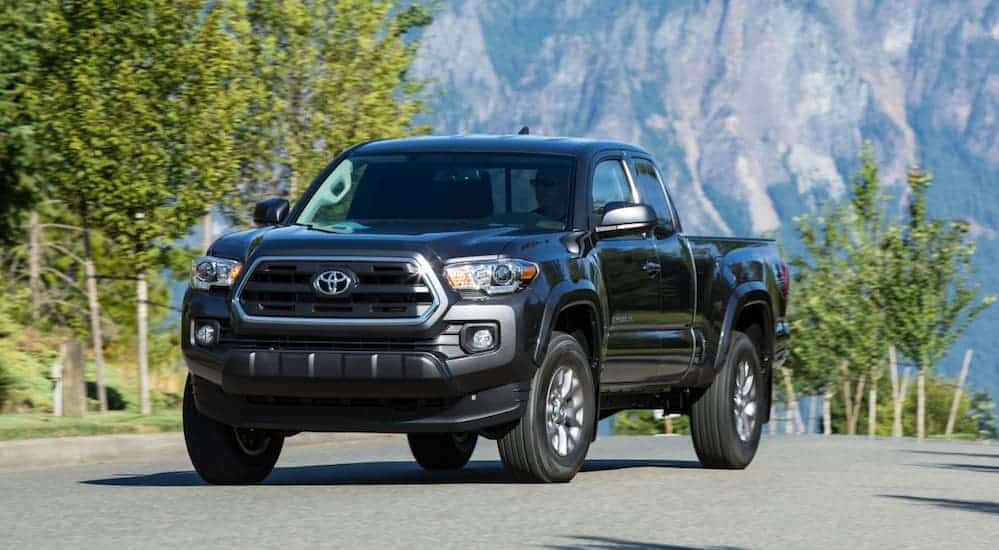 A grey 2017 Toyota Tacoma is driving on a tree lined road with mountains in the distance. 