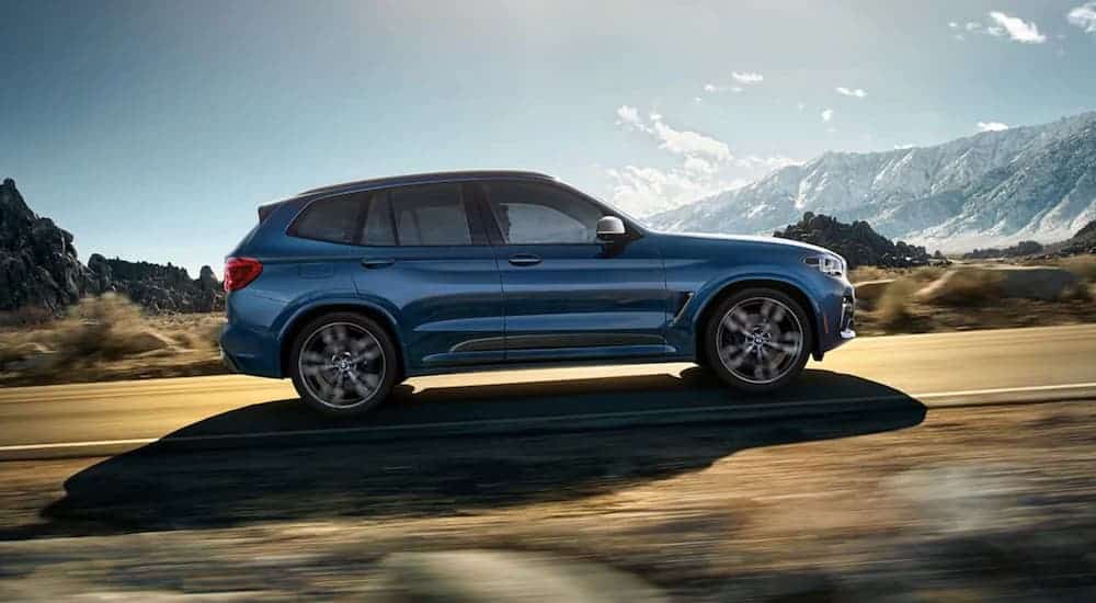 A blue 2020 BMW X3 is shown from the side driving with mountains in the distance.