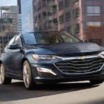 A dark blue 2020 Chevy Malibu is driving on a city street.