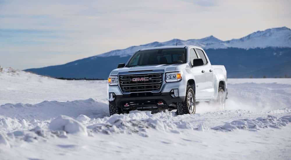 A white 2021 GMC Canyon AT4 is driving on a snow covered road with mountains in the distance. 