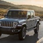 A grey 2020 Jeep Gladiator is driving on a grass lined road while the sun sets in the distance.