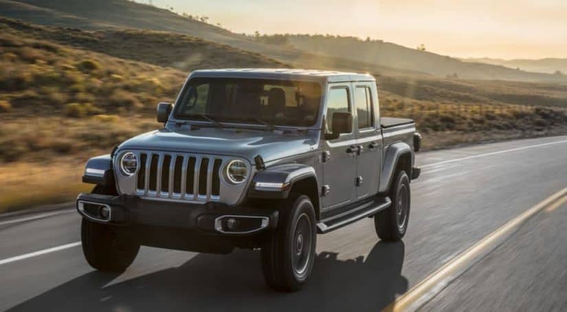 A grey 2020 Jeep Gladiator is driving on a grass lined road while the sun sets in the distance.