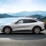 A white 2021 Ford Mustang Mach-E is driving by mountains and a lake.