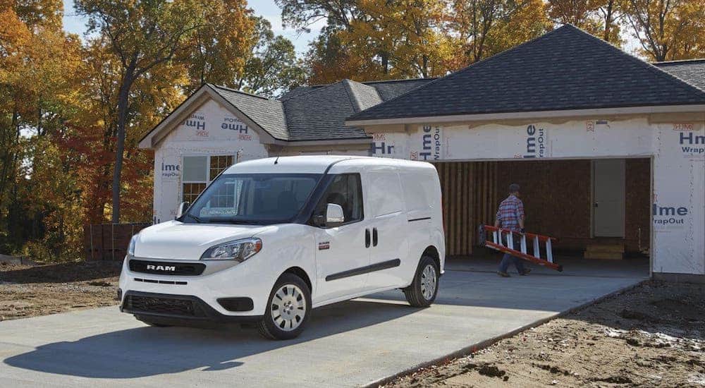 A man carrying a ladder is walking away from an unmarked white 2020 Ram ProMaster van towards a house that is under construction.