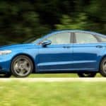 A blue 2017 Ford Fusion Sport is driving on a rural road away from a Used Car Dealerships Near Me.