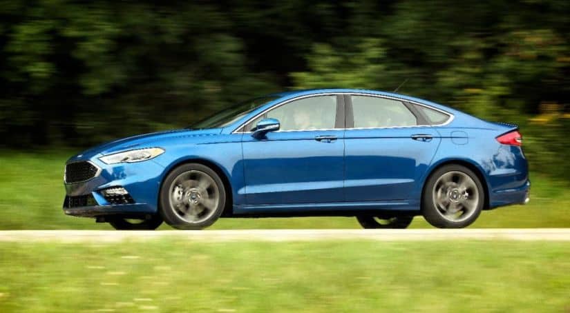 A blue 2017 Ford Fusion Sport is driving on a rural road away from a Used Car Dealerships Near Me.