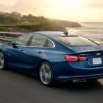 A blue 2020 Chevy Malibu is driving along the coast at sunset.