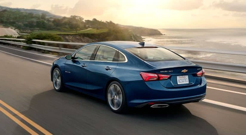 A blue 2020 Chevy Malibu is driving along the coast at sunset.