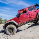 A red Jeep Wrangler Unlimited is climbing down a rock against a blue sky.