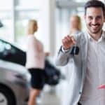 A car salesman is holding keys in front of two women shopping for used cars for sale.