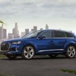 A blue 2020 Audi Q7 is parked with a city skyline in the distance.