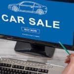 A computer screen is shown from a high angle with "car sale" on the screen, also shown are a keyboard, hands holding coffee and a pencil, a small plant.