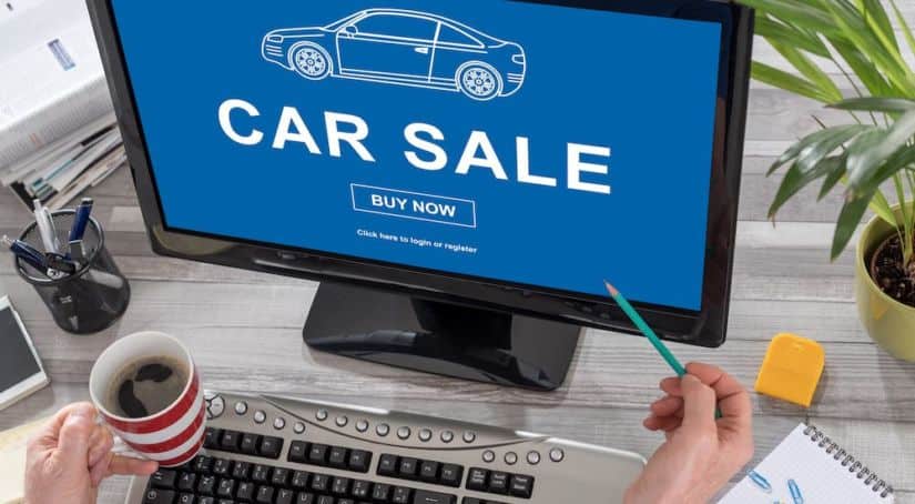A computer screen is shown from a high angle with "car sale" on the screen, also shown are a keyboard, hands holding coffee and a pencil, a small plant.