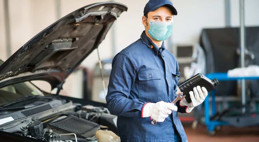 A mechanic is leaning on a car while wearing a face mask.