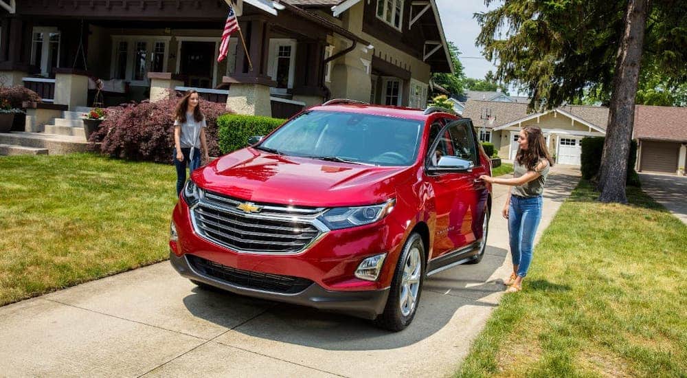 Two young women are getting into a popular used car, a red 2018 Chevy Equinox, which is parked in a driveway.