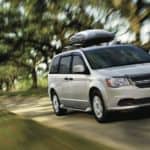 A silver 2020 Dodge Grand Caravan with a luggage compartment on top is driving on a tree-lined road.