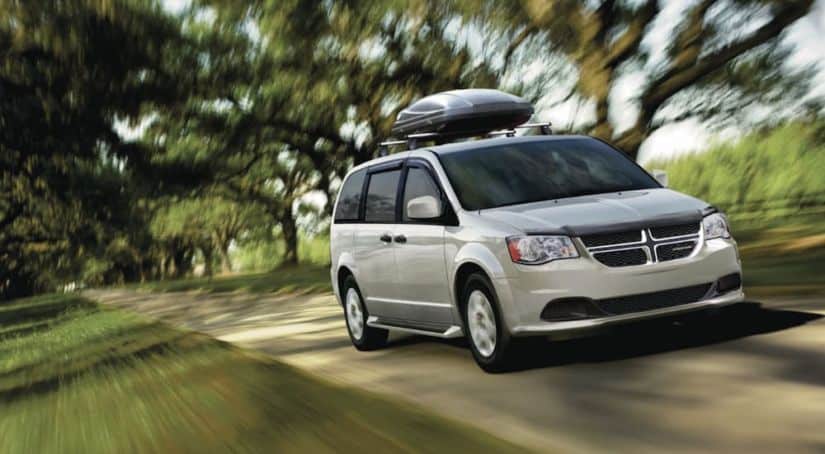 A silver 2020 Dodge Grand Caravan with a luggage compartment on top is driving on a tree-lined road.